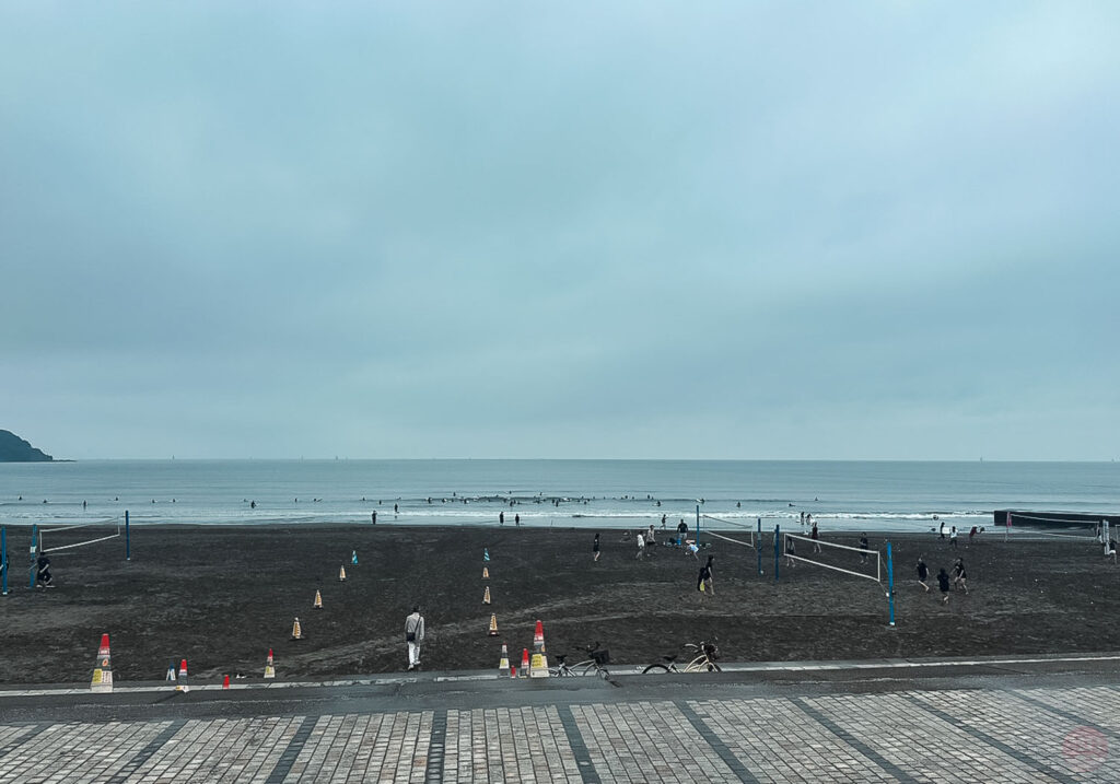 曇り空の海岸でビーチバレーを楽しむ人々と、海面に浮かぶ多くのサーファー達